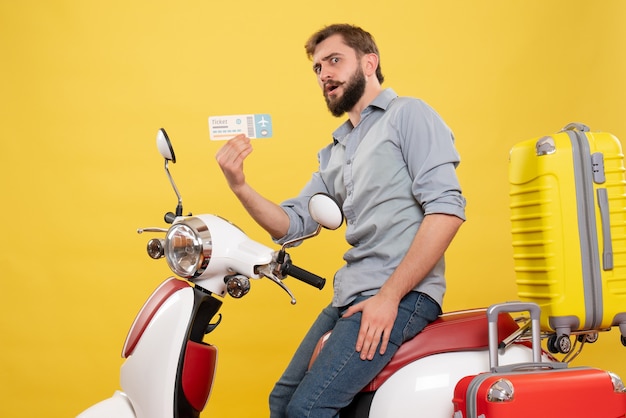 黄色のチケットを保持しているスーツケースとバイクに座っているひげを生やした若い男と旅行の概念の正面図