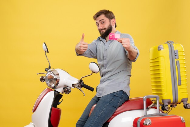 黄色の銀行カードを保持しているスーツケースとバイクに座って幸せな笑顔の若い男と旅行コンセプトの正面図