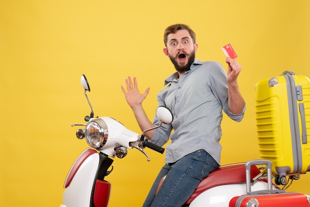Front view of travel concept with surprised emotional young man sitting on motocycle with suitcases on it holding bank card on yellow 
