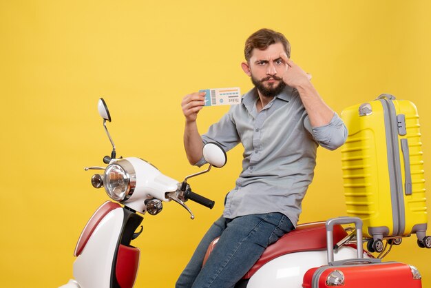 黄色のチケットを保持しているスーツケースを持ってオートバイに座っている不思議な考えの若い男と旅行コンセプトの正面