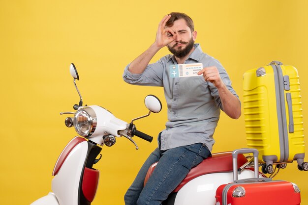 黄色のそれに眼鏡のジェスチャーを作るチケットを示す、オートバイに座っている若い自信を持ってひげを生やした男と旅行の概念の正面図