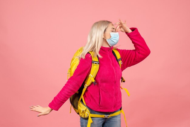 Женщина-путешественница с желтым рюкзаком в маске, держащая нос, вид спереди