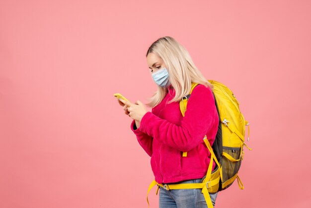 Front view traveler woman with yellow backpack wearing mask looking at phone
