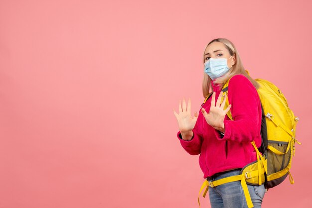 Front view traveler woman with yellow backpack wearing mask making stop sign