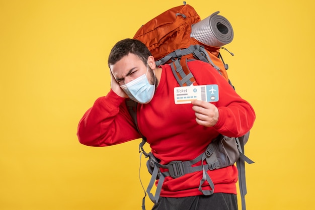 Free photo front view of traveller guy wearing medical mask with backpack showing ticket and feeling nervous on yellow background