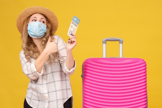 Front view of travelling girl wearing mask showing ticket and standing near her pink bag making ok gesture on yellow 