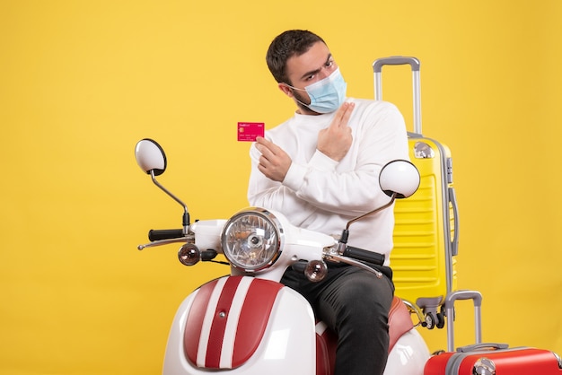 Front view of trip concept with confident young guy in medical mask sitting on motorcycle with yellow suitcase on it and holding bank card