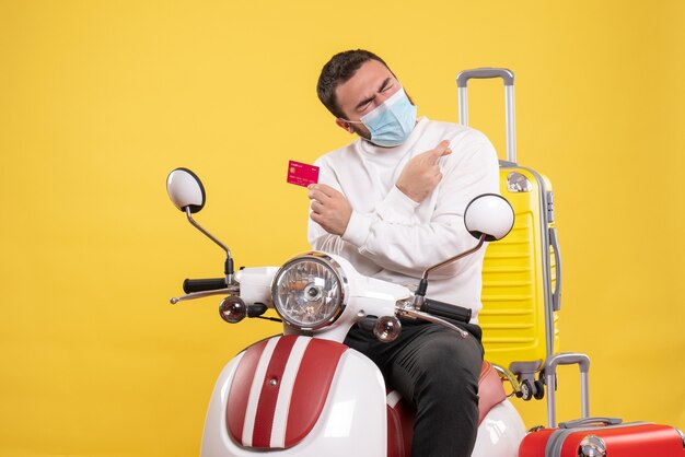 Front view of trip concept with young hopeful guy in medical mask sitting on motorcycle with yellow suitcase on it and holding bank card