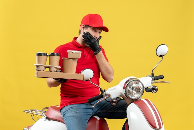 Front view of troubled courier man wearing red blouse and hat gloves in medical mask delivering order sitting on scooter holding orders suffering from toothache