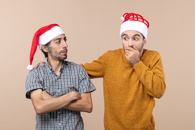 Front view two amazed guys with santa hats one putting his hand on the others shoulder on beige isolated background