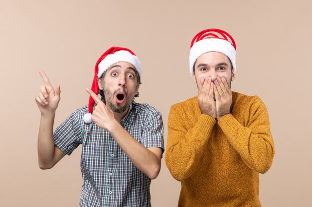 Front view two amazed guys with santa hats one showing something the other covering face with his hands on beige isolated background