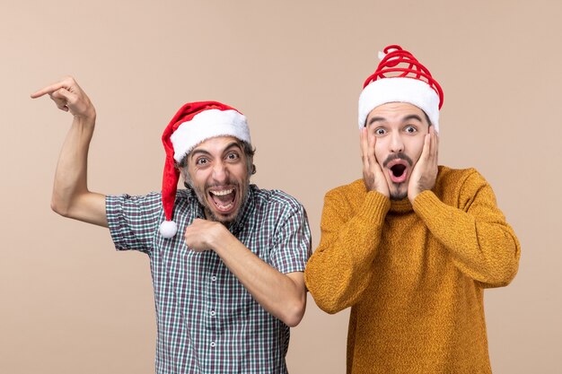 Front view two amazed guys with santa hats one showing something and the other shocking on beige isolated background