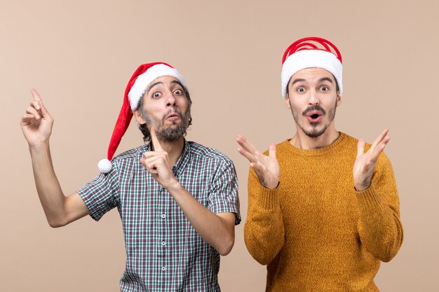 Front view two amazed guys with santa hats one showing something the other shocking on isolated background