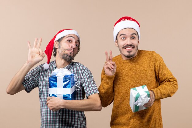 Front view two blissful guys one making okey sign and the other victory sign and holding christmas presents on beige isolated background