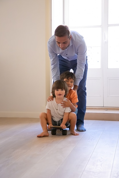 Front view of two boys riding on skateboard at home. Caucasian attractive father pushing his lovely sons to back and playing with kids. Childhood, game activity and weekend concept