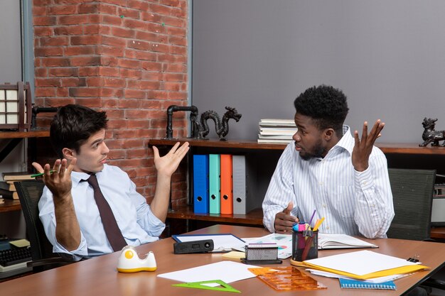 Front view two businessmen in formal wear sitting at table with office stuffs discussing