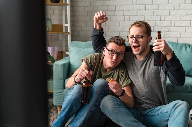 Front view of two cheerful male friends watching sports on tv and having beer