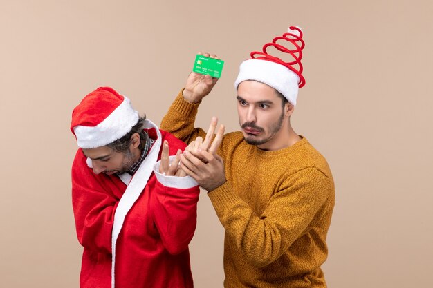 Front view two confused guys one with santa coat looking at floor and the other holding card on beige isolated background