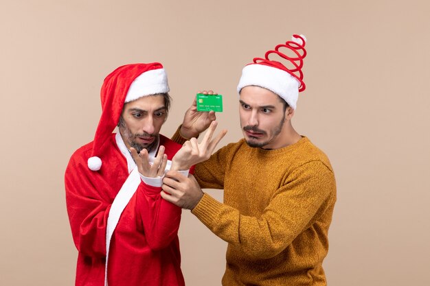 Front view two confused guys one with santa coat and the other holding card on beige isolated background