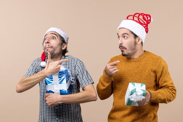 Вид спереди два запутанных парня указывают пальцем, показывая что-то и держат рождественские подарки на бежевом изолированном фоне