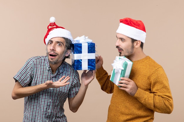 Front view two confused guys wearing santa hats and checking their christmas presents on beige isolated background