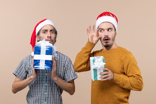 Бесплатное фото Вид спереди двух сбитых с толку парней в шляпах санта-клауса с рождественскими подарками, один из которых приложил руку к скуле