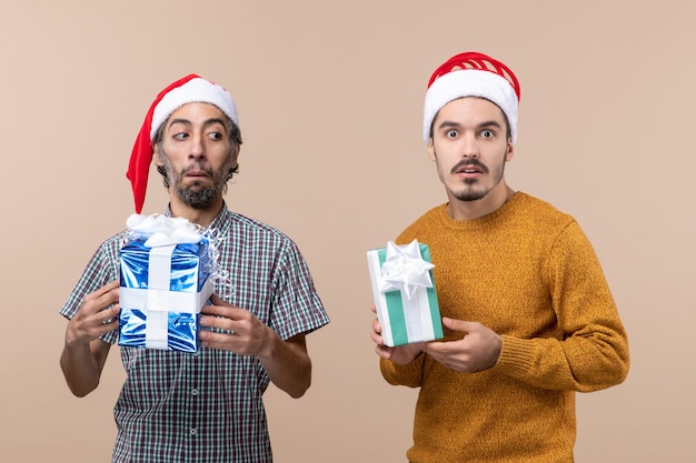 Vista frontale di due ragazzi confusi che indossano cappelli di babbo natale e tengono in mano regali di natale