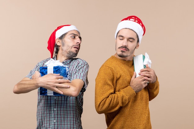 Vista frontale due ragazzi confusi che indossano cappelli di babbo natale e tengono stretti i loro regali su sfondo beige isolato