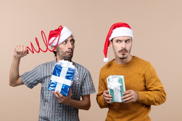 Front view two confused guys with santa hats and christmas presents trying to concentrate on beige isolated background