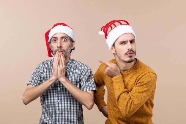 正面図サンタの帽子をかぶった2人の好奇心旺盛な男が、片方は彼の顎に手を置き、もう一方の人差し指はベージュの孤立した背景に彼の友人を示しています