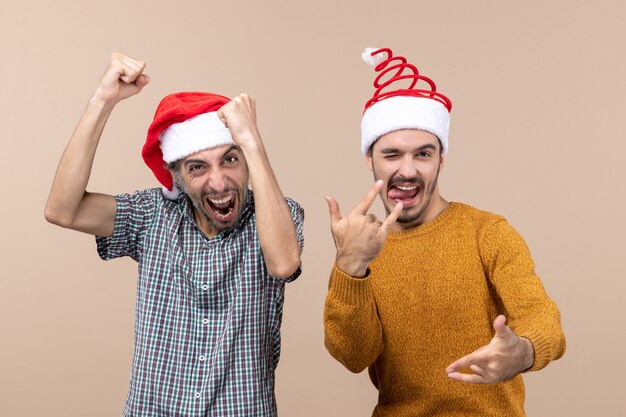 Вид спереди двух приподнятых парней в шляпах санта-клауса, показывающих свое счастье на бежевом изолированном фоне