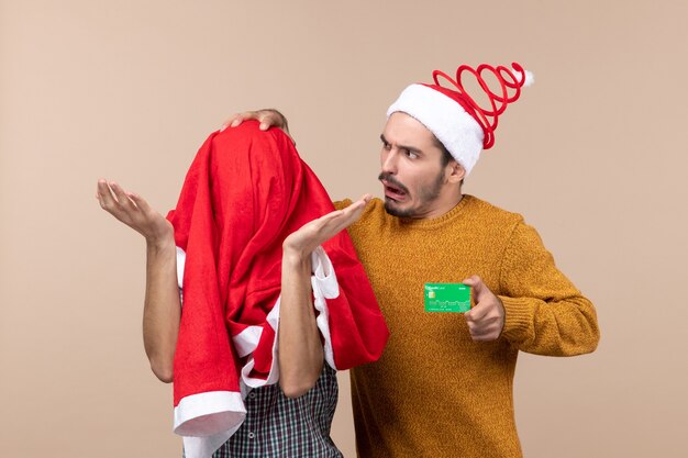 그의 머리를 만지고 다른 베이지 색 격리 된 배경에 산타 코트와 그의 머리를 덮고있는 동안 전면보기 두 친구 하나는 신용 카드를 들고
