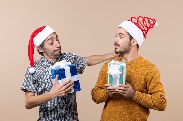 Front view two friends wearing santa hats and holding presents on beige isolated background