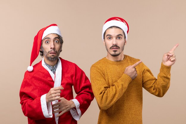 Front view two friends with santa hats one confusing and the other showing direction on beige isolated background