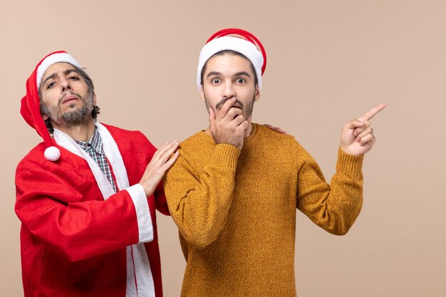 산타 모자 하나는 다른 어깨를 잡고 다른 하나는 베이지 색 격리 된 배경에 방향을 보여주는 전면보기 두 친구
