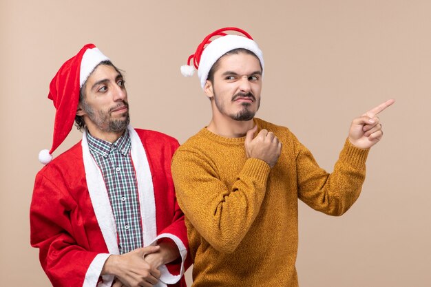 Front view two friends with santa hats one looking at the other and the other showing direction on beige isolated background