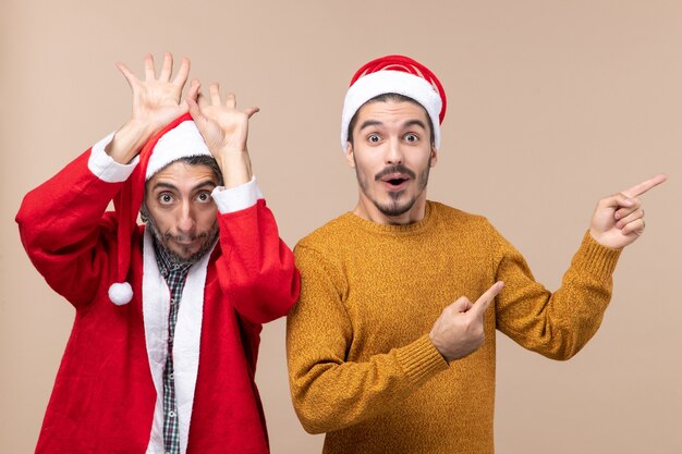 Front view two friends with santa hats one raising his hands to his head and the other showing direction on beige isolated background