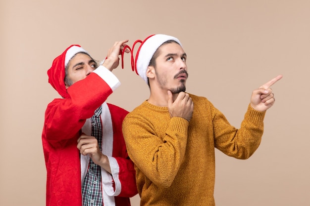 Front view two friends with santa hats one trying to holding hats pompom and the other showing direction on beige isolated background