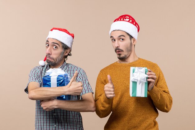 Front view two gratified guys making thumb up sign and holding christmas presents on beige isolated background