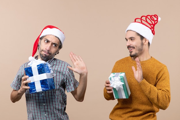 Вид спереди двух обиженных и счастливых парней в шляпах санта-клауса и рождественских подарков на бежевом изолированном фоне