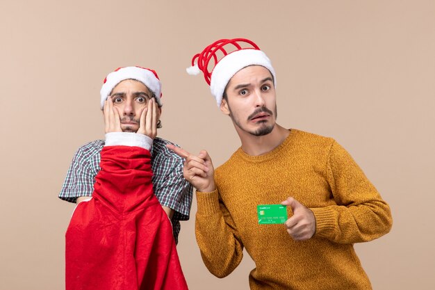 Front view two guys one with santa coat putting hands on his face and the other with credit card on beige isolated background