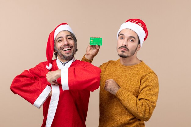 Front view two guys one with santa coat smiling and the other with credit card looking at camera on beige isolated background