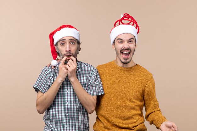 Front view two guys with santa hats one depressing and the other laughing on beige isolated background