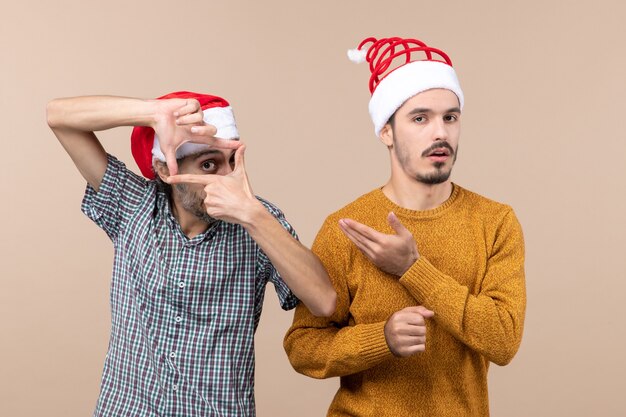 Front view two guys with santa hats one making camera sign with his hands and the other showing him on beige isolated background