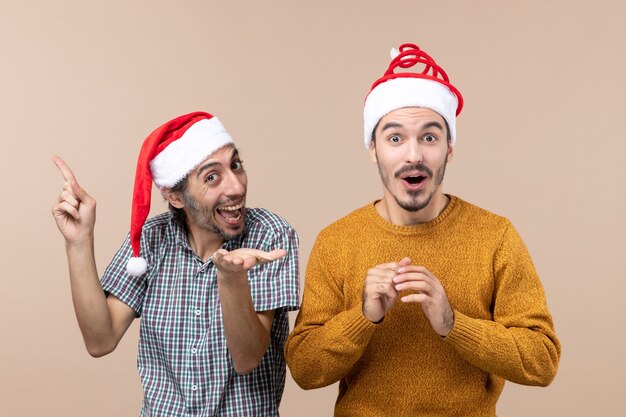 Front view two guys with santa hats one showing something the other surprising on beige isolated background