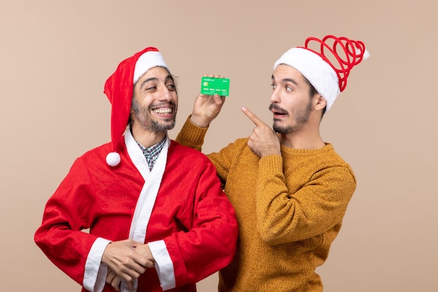 Front view two happy guys one with santa coat and the other with credit card looking at it on beige isolated background