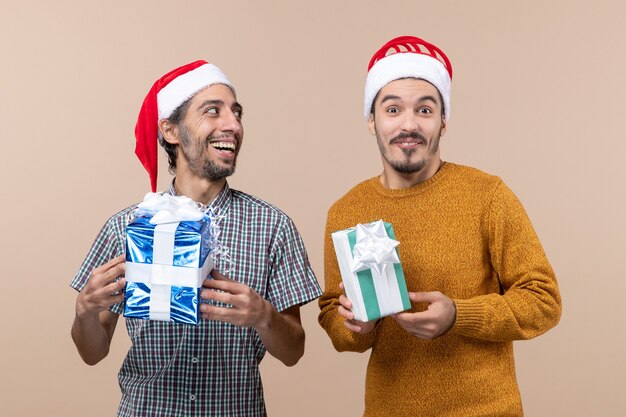 Front view two happy guys wearing santa hats and holding christmas presents on beige isolated background