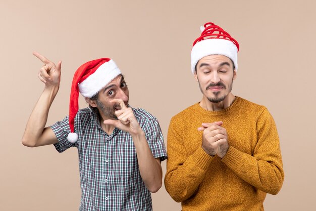 Front view two happy guys with santa hats one showing something the other closing his eyes and holding his hands together on beige isolated background