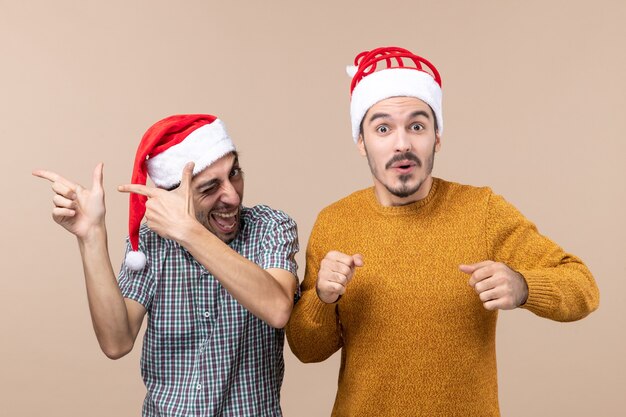 Front view two happy guys with santa hats one with blinked eye and the other with prying eyes on beige isolated background