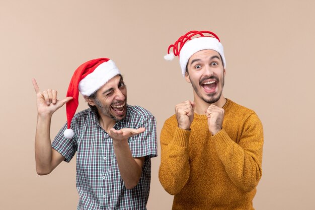 Front view two happy guys with santa hats one with blinked eye the other with punch fist on beige isolated background